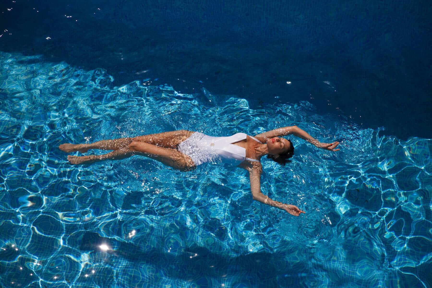 Woman gracefully floating in a turquoise blue pool, dressed in an elegant white one piece bathing suit